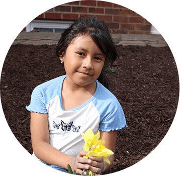 A young girl with long dark hair sits outdoors on brown mulch, holding a yellow flower. She wears a light blue and white shirt with butterfly designs and looks toward the camera with a slight smile.