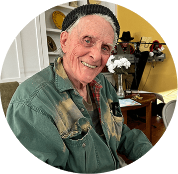 An older man with a gray beard, wearing a green shirt and black knit hat, smiles while sitting indoors. Shelves with plates and white flowers are visible in the background.