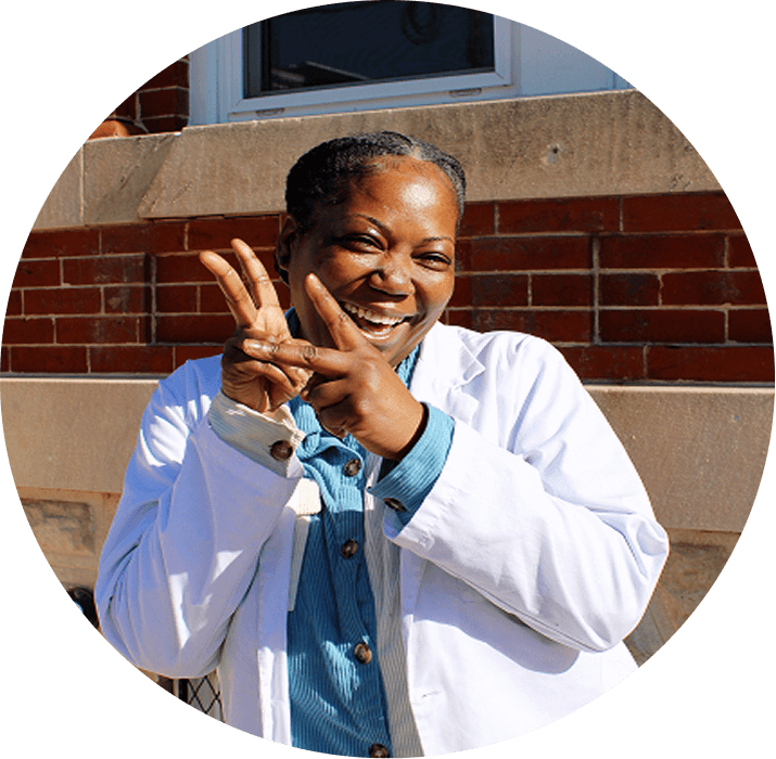 A smiling woman in a white lab coat and blue shirt stands outside in front of a brick wall, making peace signs with both hands. For more information or questions, contact AHC.