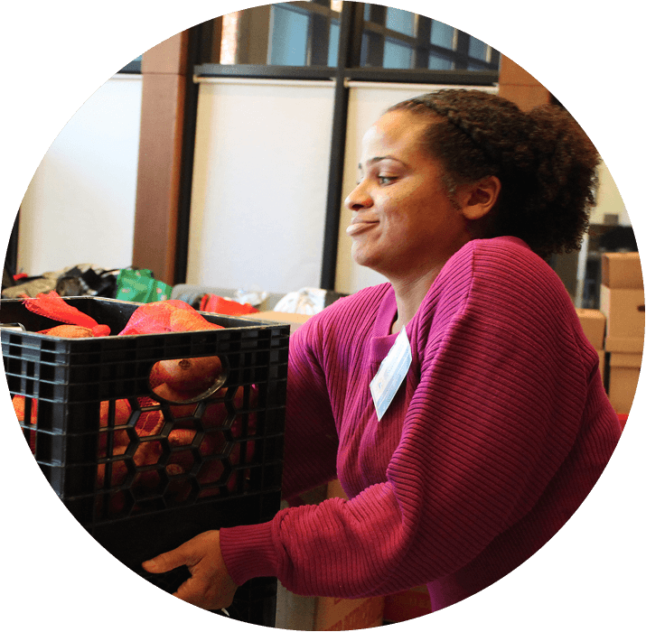 A woman wearing a magenta sweater smiles while lifting a black crate filled with red apples in an indoor setting. For more information, contact AHC.