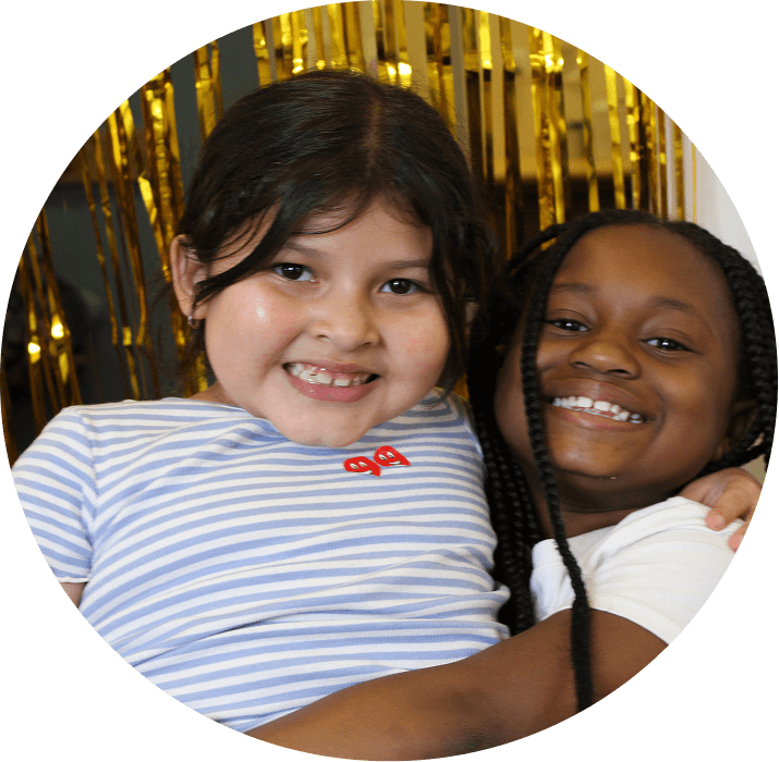 Two young children smile and hug each other in front of a gold tinsel backdrop. One wears a striped shirt, while the other has braided hair and a white shirt. For more details, contact AHC.