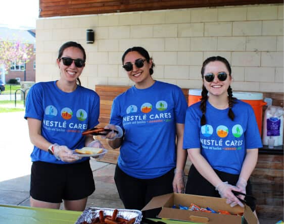 Three women in matching blue Nestlé Cares shirts and sunglasses smile while serving food outdoors. Supporting Affordable Homes & Communities, they distribute hot dogs and snacks with gloves at a lively community event.