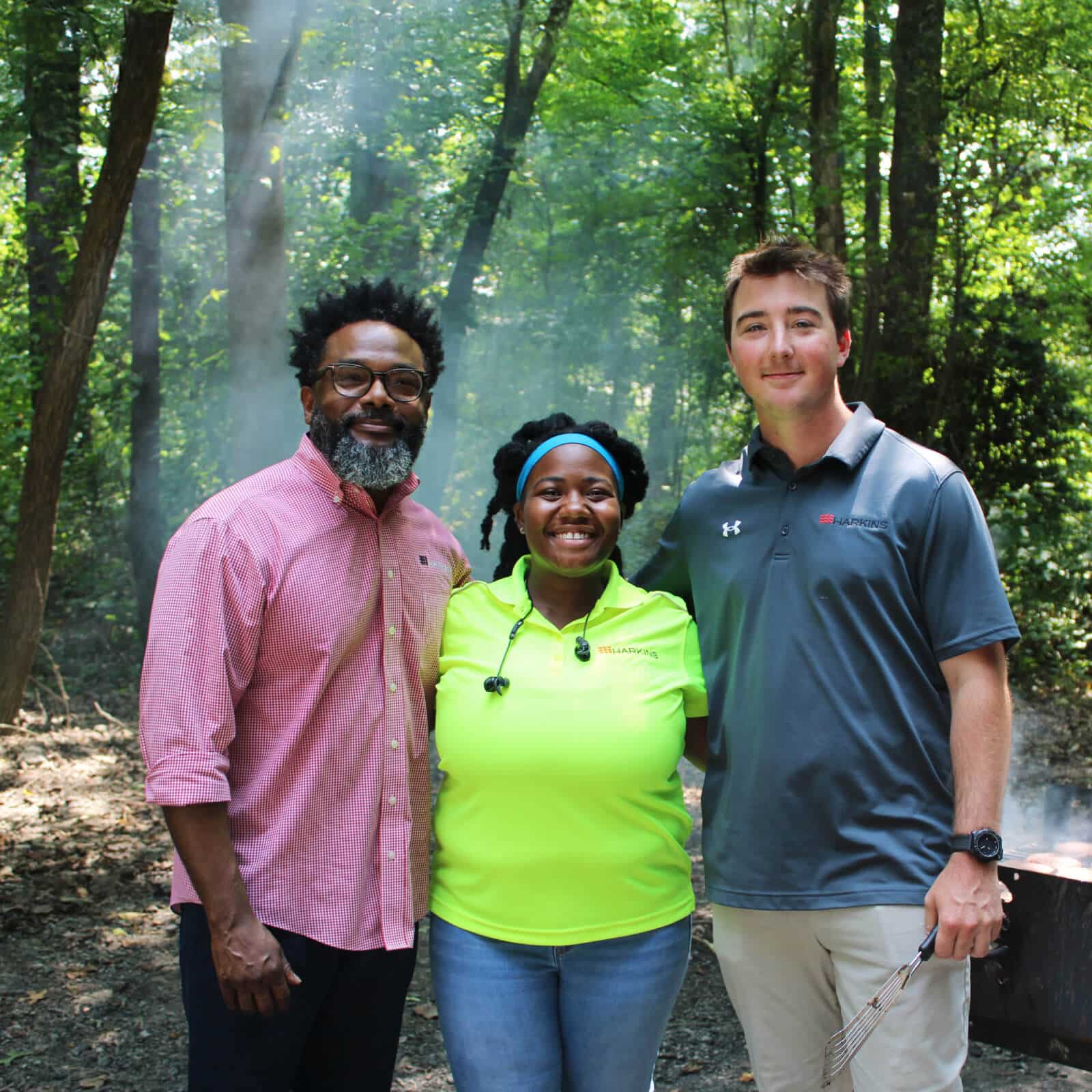 Three adults stand together smiling in a sunlit wooded area, showcasing the spirit of Affordable Homes & Communities. The person on the left wears glasses and a pink shirt, the center person is in neon yellow, and the right person wears a gray polo.