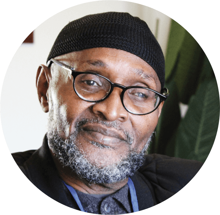 A middle-aged man with a gray beard, black glasses, and a black knit cap smiles gently at the camera. He is wearing a dark shirt and jacket, with the AHC Impact Report 2024 open beside him and a blurred green plant in the background.