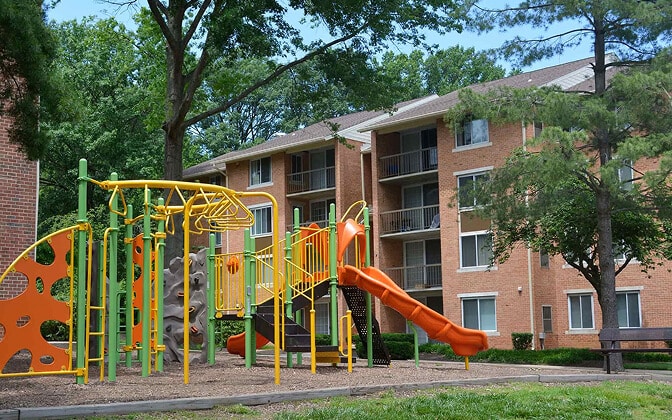 A colorful playground with slides and climbing structures sits in front of a brick apartment building, surrounded by trees and greenery.