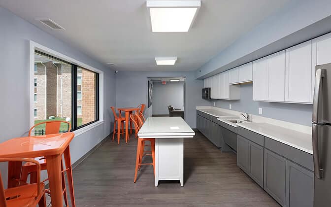 Modern communal kitchen with gray lower cabinets, white upper cabinets, a central island, stainless steel appliances, and orange chairs around small tables by a large window. The room has gray floors and light blue walls.