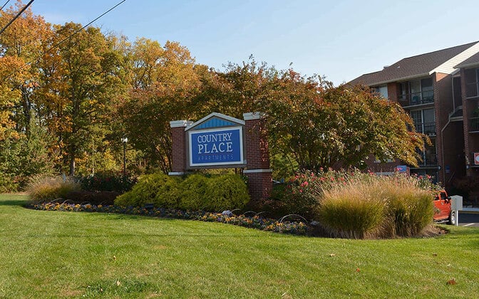 A brick sign reads Country Place Apartments amid landscaped bushes and flowers, with apartment buildings and trees in the background on a sunny day.