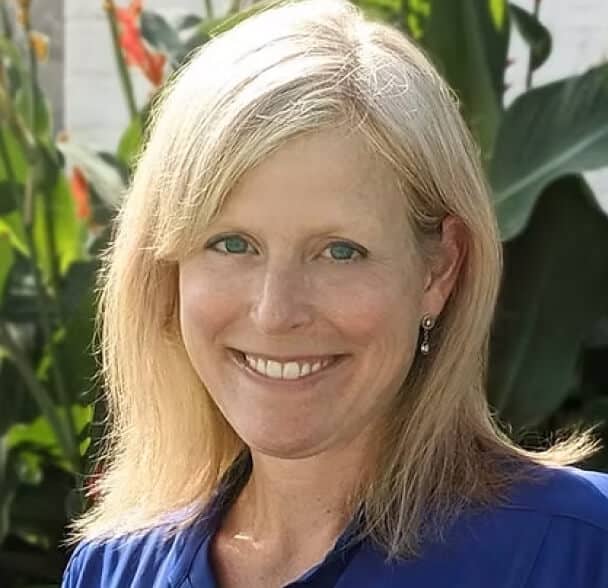 A smiling woman with straight blonde hair and blue eyes, wearing a blue top and drop earrings, stands outdoors in front of green plants with orange flowers.