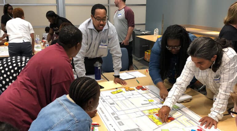 A group of people gathers around a table, collaborating and discussing Building Community With AHC Welcome Baskets as they point to a large map or planning board covered with colorful markers and notes in a bright, modern meeting room.