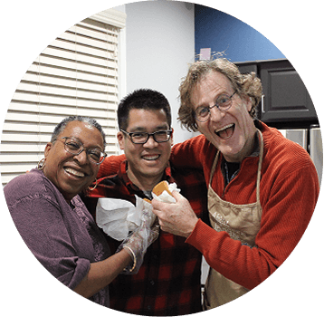 Three smiling adults stand close together in a kitchen, one woman in a purple shirt, a man in glasses and a red plaid shirt, and another man in a red sweater and apron, holding food and posing happily.