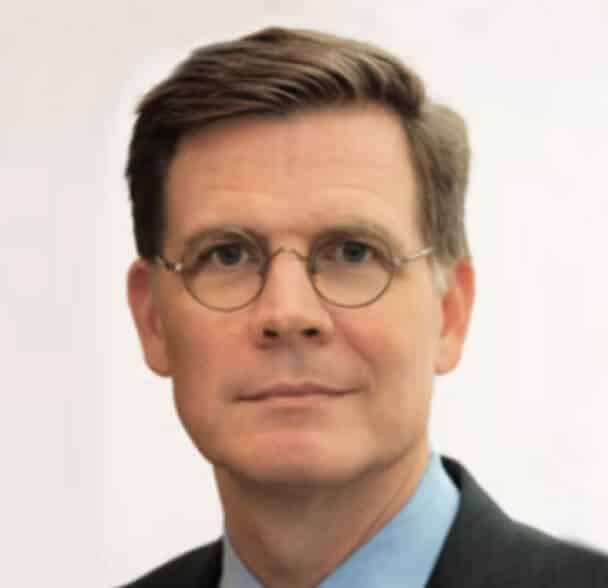 A man with short brown hair, wearing round eyeglasses, a blue shirt, and a dark jacket, poses against a plain, light background.
