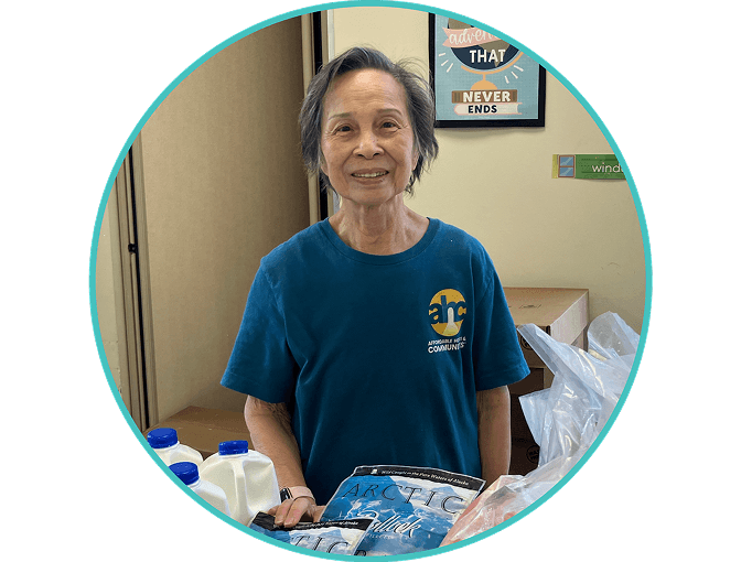 An older woman with short gray hair smiles while standing behind a table with milk cartons and frozen food. She wears a blue t-shirt with a yellow logo. Behind her, motivational posters are visible on the wall.