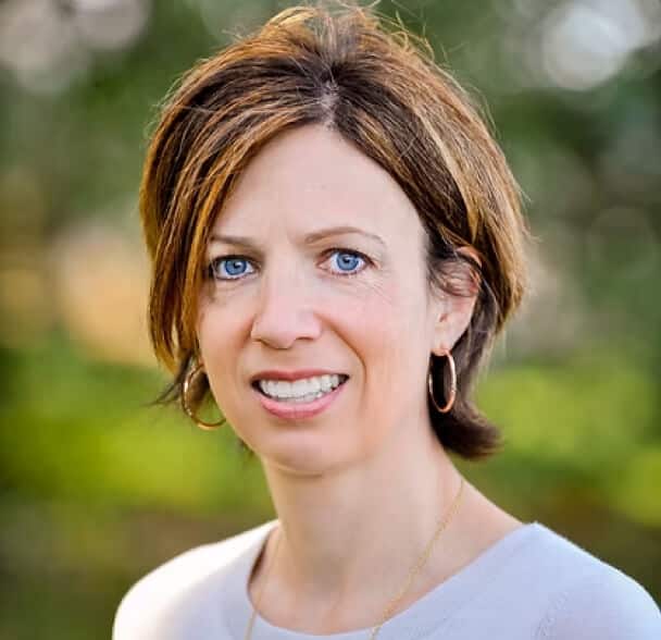 A woman with short brown hair and blue eyes, wearing hoop earrings and a light top, stands outdoors with a blurred green background, smiling softly at the camera.