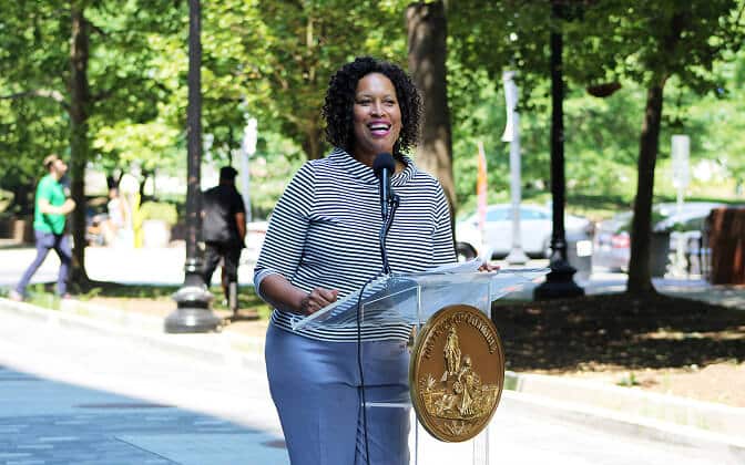 A woman stands outdoors at a clear podium with a gold seal, smiling and speaking into a microphone. Trees, parked cars, and people walking are visible in the background.