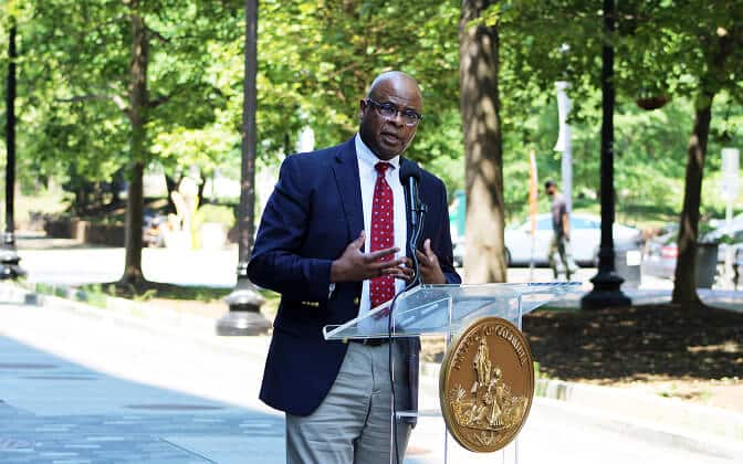 A man wearing glasses, a suit, and a red polka-dot tie speaks at an outdoor podium with a gold city seal, surrounded by trees and sunlight.