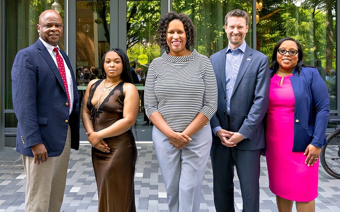Five people stand side by side outdoors, posing for a group photo. They are dressed in formal and semi-formal attire, smiling and standing in front of a glass building with greenery reflected in the windows.