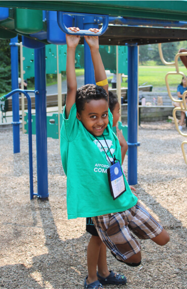 A smiling child in a green shirt hangs from blue monkey bars at a playground, with mulch on the ground and playground equipment in the background.