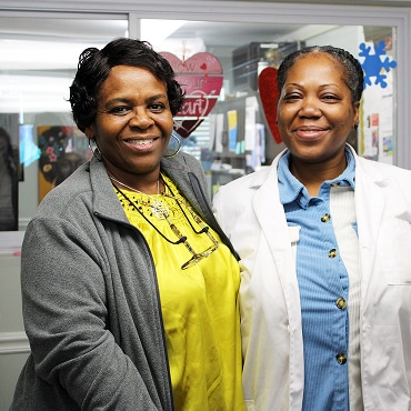Two women smiling and standing side by side indoors in front of a decorated window with heart and snowflake cutouts, celebrating the spirit of Affordable Homes & Communities. One wears a yellow top and gray jacket; the other, a blue shirt and white coat.