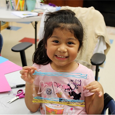 A young girl smiles and holds up a clear sheet decorated with colorful drawings of hearts, waves, and shapes—her creativity shining in a space supported by Affordable Homes & Communities. Art supplies and papers are on the table in front of her.