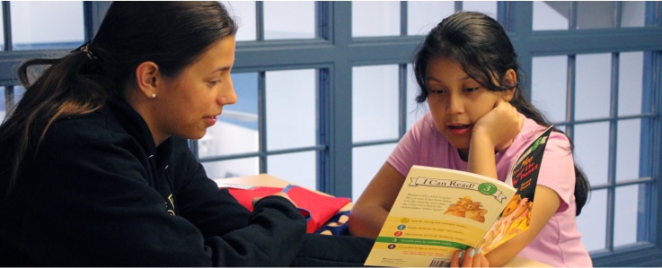 Two girls sit at a table in a room with blue-framed windows in an Affordable Homes & Communities setting; one listens attentively while the other, wearing a pink shirt, reads aloud from a children’s book titled I Can Read!.