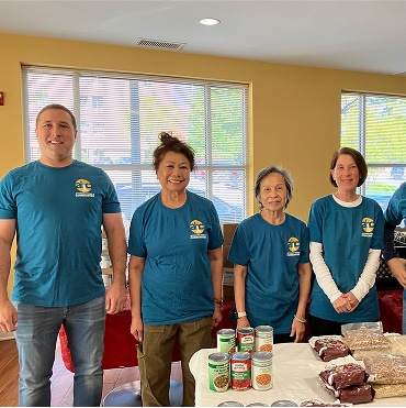 Five people wearing matching blue shirts stand behind a table with canned goods and packaged food in a brightly lit room. Smiling, they appear to be volunteering to support Affordable Homes & Communities.