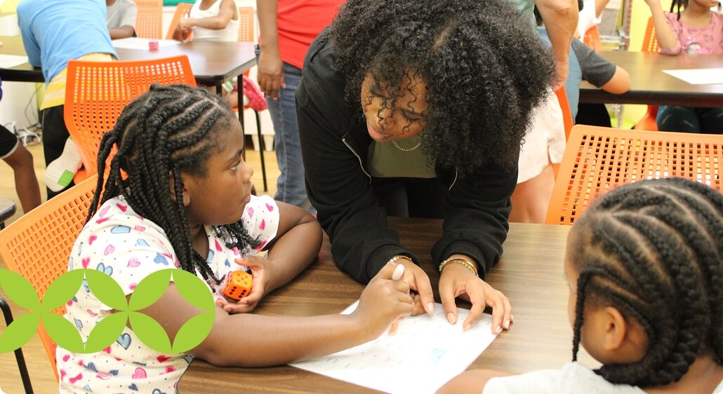 A woman leans over a table, helping two young girls with braided hair as they work on a worksheet in a classroom setting, echoing the dedication highlighted in the AHC Impact Report 2024. Other children and adults are visible in the background.