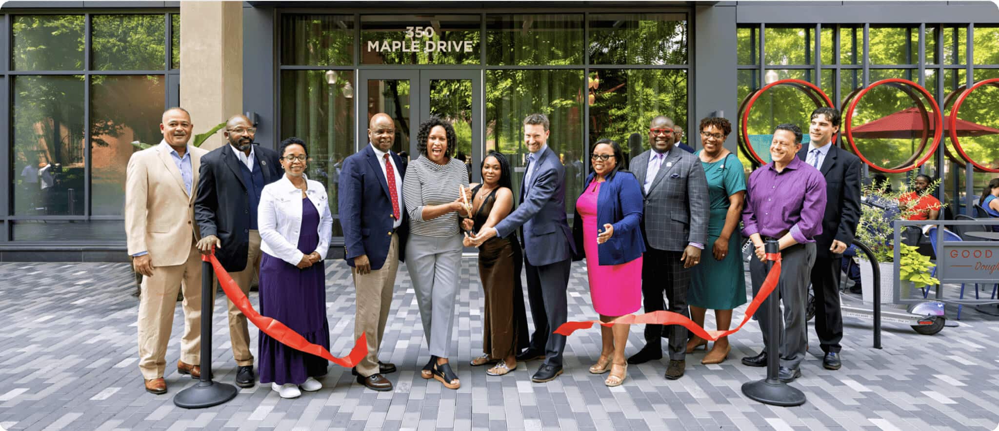 A group of people smiles and poses together in front of a building at 350 Maple Drive, holding a red ribbon in a ribbon-cutting ceremony featured in the AHC Impact Report 2024. Some hold scissors, marking the opening event.
