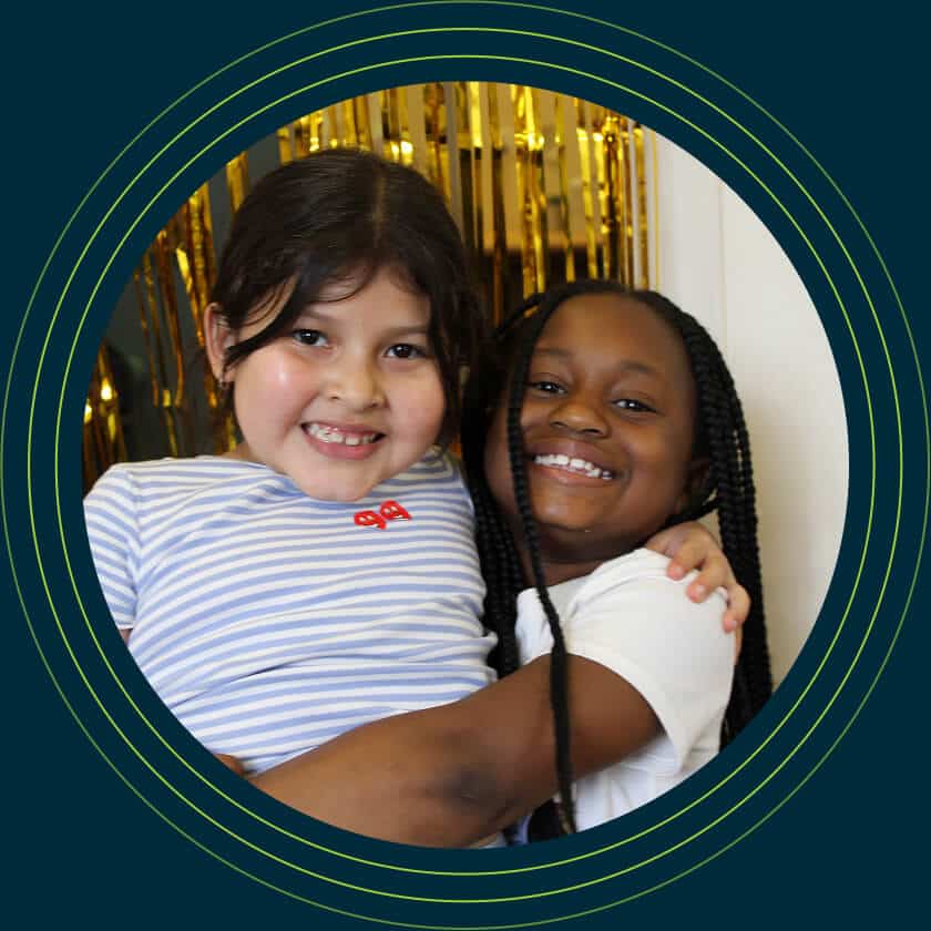 Two smiling young girls, one with long dark hair in a striped shirt and one with braided hair in a white shirt, hug each other in front of a gold tinsel backdrop, framed by green concentric circles, celebrating the AHC Impact Report 2024.