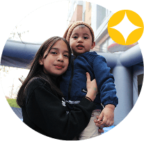 A young girl holds a small child outside, both looking at the camera. The child wears a tan beanie and blue jacket. A yellow star-shaped icon in the top right hints at their feature in the AHC Impact Report 2024.