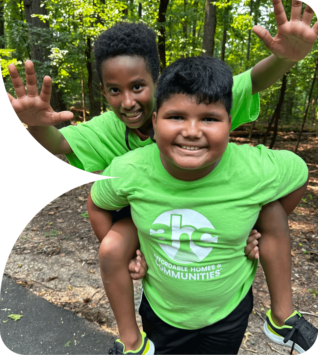 Two smiling boys outdoors in bright green “Affordable Homes & Communities” T-shirts enjoy a piggyback ride on a wooded path, both waving at the camera. Their joyful moment captures the spirit of our AHC Impact Report 2024.