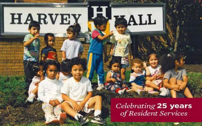 A group of young children sit and stand together smiling in front of a Harvey Hall sign. A banner in the corner reads: “Celebrating 25 years of Resident Services.”.
