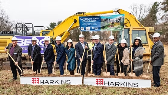 A group of people in hard hats and formal attire hold shovels at a Habitat for Humanity breaking ground ceremony in front of an excavator and a sign reading “Randolph Road Community.” Two banners display the name Harkins.
