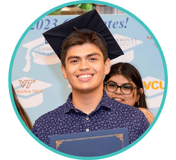A young man in a graduation cap smiles while holding a certificate, with a blue and white banner and other people in the background.