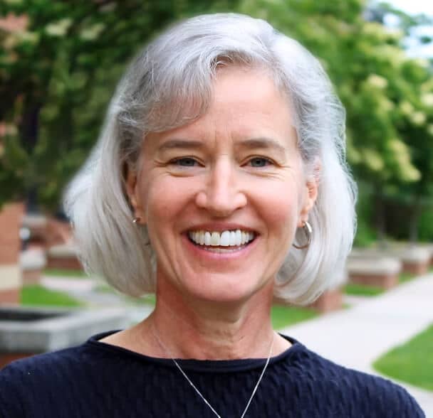 A smiling older woman with short gray hair and hoop earrings stands outdoors, wearing a navy textured top and a silver necklace. Green trees and a pathway are blurred in the background.