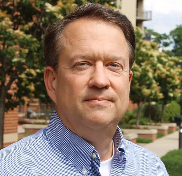 A middle-aged man with short brown hair, wearing a blue checkered shirt, stands outdoors in front of trees and a brick building, looking directly at the camera with a neutral expression.