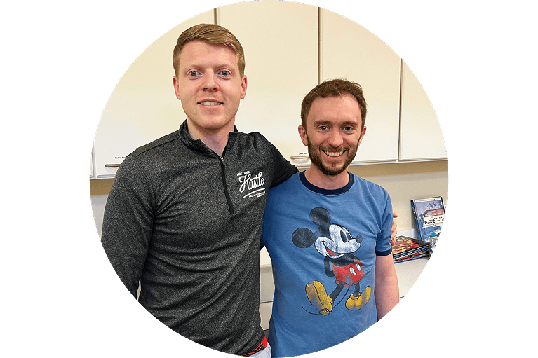 Two men smiling and standing side by side in a kitchen. One wears a dark zip-up shirt with a logo, the other wears a blue Mickey Mouse T-shirt. Some board games are visible on a counter behind them.