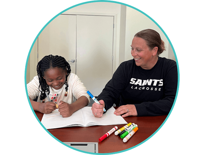 A smiling adult and a young girl sit at a table, working together in a notebook. Colorful markers are on the table as they both write and laugh, enjoying their activity.