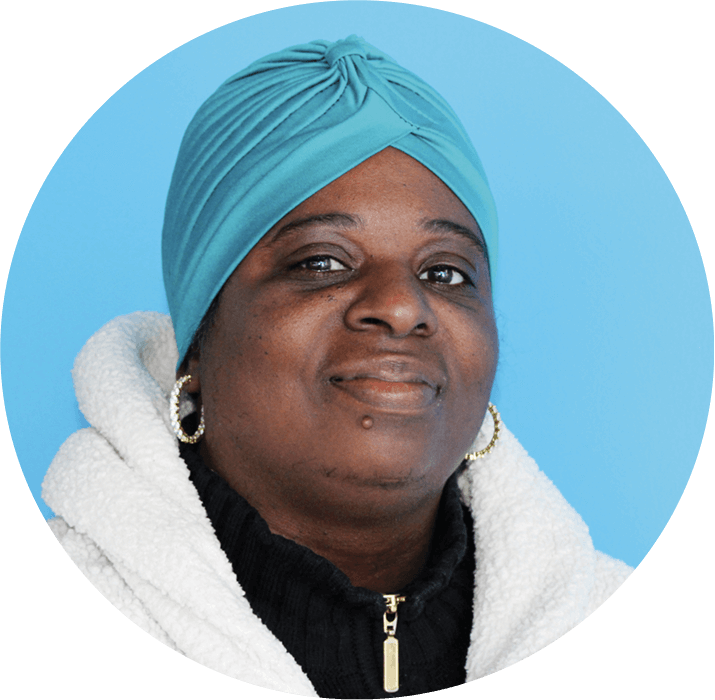 A woman wearing a turquoise headwrap, white fleece jacket, and black top with a gold zipper poses in front of a solid blue background. Featured in the AHC Impact Report 2024, she smiles slightly and wears hoop earrings.