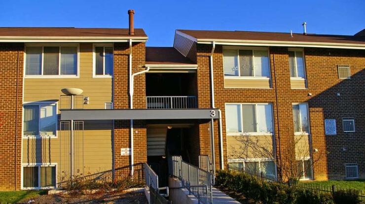 A two-story brick and tan apartment building with a central entrance and ramp, large windows, and a flat roof, pictured in bright sunlight with a clear blue sky.
