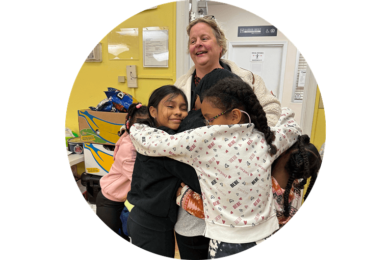 Three young girls are hugging each other and smiling in a classroom, with an adult woman standing behind them, also smiling. The setting appears warm and cheerful.