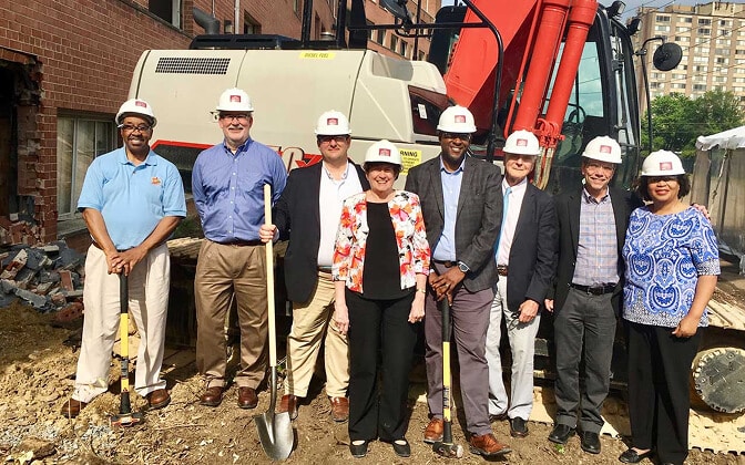 Eight people wearing hard hats stand in front of construction equipment at a building site, some holding shovels, posing and smiling for a groundbreaking ceremony photo.
