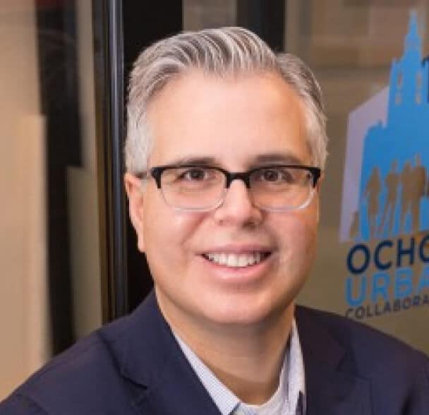 A smiling person with short gray hair and glasses wearing a dark blazer stands in front of a window with a blue urban-themed logo and text in the background.