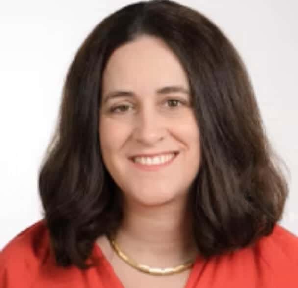 A woman with shoulder-length dark brown hair, wearing a red top and a gold necklace, smiles at the camera against a plain light background.