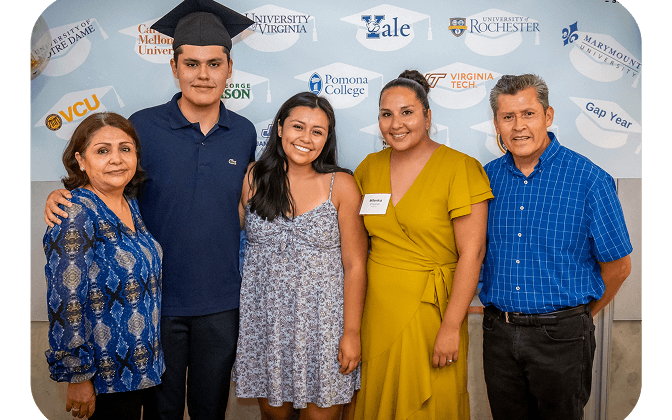 Five people stand smiling in front of a wall decorated with various university logos. One young man wears a graduation cap, flanked by two women and an older man and woman. They appear to be celebrating a graduation.