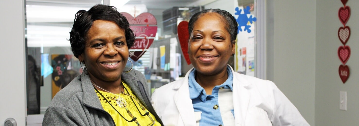 Two women smile and stand close together indoors, one in a gray jacket and yellow shirt, the other in a white medical coat. Heart-shaped decorations adorn the walls, creating a warm scene for the Affordable Homes & Communities Media Center.