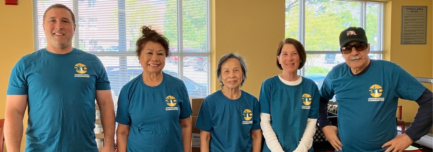 Five adults stand indoors, smiling at the camera in matching blue shirts with the Affordable Homes & Communities Media Center logo. Sunlight pours through large windows behind them, with tables and boxes visible in the background.