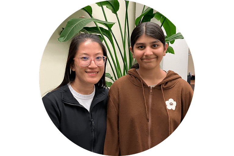 Two people stand side by side indoors, smiling at the camera. One wears glasses and a black jacket, the other wears a brown hoodie with a flower pin. Green leafy plants are in the background.