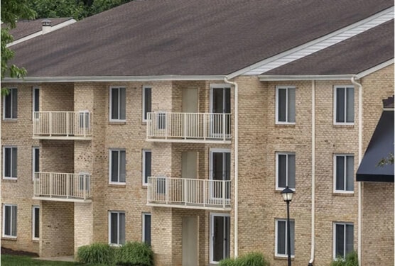 A beige brick apartment building with white railings on small balconies, multiple windows, and a sloped dark roof—an example of the quality housing seen in 9 Ways AHC Boosts Economic Mobility. Green bushes adorn the ground level near a welcoming entrance.