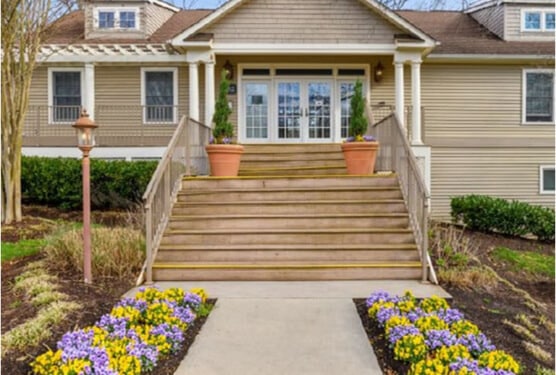 A wide staircase with railings leads up to the front entrance of a beige house with large windows—one of the 9 Ways AHC Boosts Economic Mobility is by creating welcoming, vibrant communities like this, complete with colorful flower beds and potted plants.