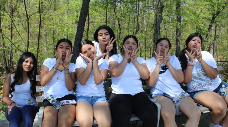 Seven young women sit on a bench outdoors, all in white tops and making playful hand gestures and faces. Trees with lush green leaves brighten the background, capturing the cheerful vibe of Nestlé Partners with AHC for NesFest.